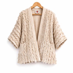 May include: A beige open-front jacket with short sleeves and two front pockets. The jacket has a textured, fringed design and is hanging on a wooden hanger. The label reads "LHASA".