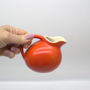 Könnte beinhalten: Eine kleine, runde, orangefarbene Keramik-Sahnekännchen mit Henkel und weißem Innenraum. Das Sahnekännchen wird von einer Hand mit lackierten Nägeln gehalten. Das Sahnekännchen ist im Vintage-Stil gehalten und wird wahrscheinlich zum Servieren von Sahne oder Milch verwendet.