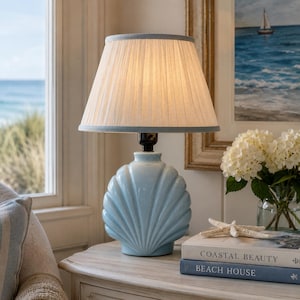 May include: A table lamp with a light blue ceramic base shaped like a seashell and a pleated off-white fabric lampshade. The lamp sits atop a stack of two books titled Coastal Beauty and Beach House, accompanied by a white starfish ornament.