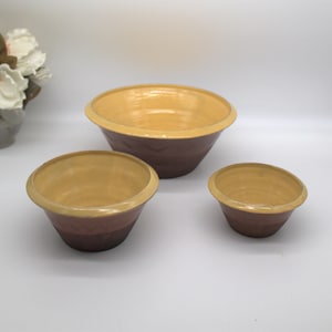 May include: A set of three ceramic mixing bowls. The bowls have a two-tone design, with a brown exterior and a yellow interior. The rims are also yellow. The bowls are nested, with the largest bowl in the back and the smallest in the front.