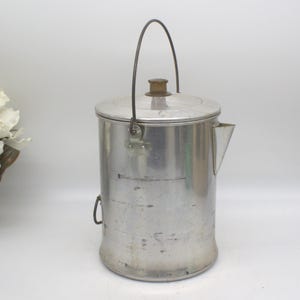 Puede incluir: Una cafetera vintage de aluminio con tapa, boquilla y asa de alambre. La cafetera tiene un cuerpo cilíndrico con una base ligeramente ensanchada. El asa está sujeta a la tapa con un pomo de madera. La cafetera está diseñada para acampar o uso al aire libre.