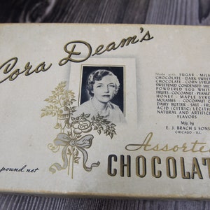 Puede incluir: Caja de bombones surtidos Cora Deam's vintage. La caja color crema presenta un retrato de una mujer, detalles florales dorados y el texto "Assorted Chocolates". La caja está etiquetada "One pound net."
