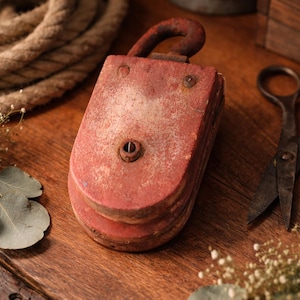 Könnte beinhalten: Eine verwitterte, rot lackierte Holzrolle mit einem rostigen Metallhaken. Die Rolle liegt auf einer Holzoberfläche, mit einem Seil und einer Vintage-Schere. Das antike nautische Werkzeug weist Gebrauchsspuren auf.