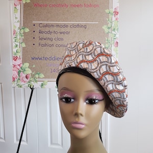 May include: A patterned beret with a black leather brim is displayed on a mannequin. The beret features a white background with orange and tan circular designs. A sign in the background reads "TEDDIES Afrique" and lists services like custom clothing and sewing classes.