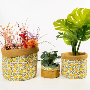 Puede incluir: Tres macetas de tela con un estampado floral amarillo y ribete de yute. Las macetas contienen flores y vegetación coloridas. La maceta más grande contiene una gran planta verde con hojas divididas.