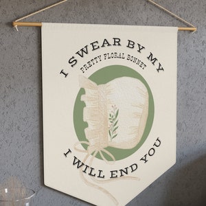 Peut inclure: Une bannière en tissu beige avec le texte "I swear by my pretty floral bonnet I will end you" en lettres noires. Un cercle vert avec un bonnet blanc à broderie florale est au centre de la bannière.
