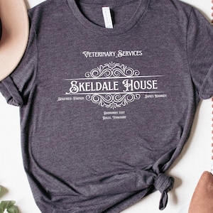 May include: A dark gray t-shirt with a white graphic design that says "Veterinary Services Skeldale House" with a decorative flourish. The graphic also includes the text "James Herriot" and "Darrowby, Yorkshire".
