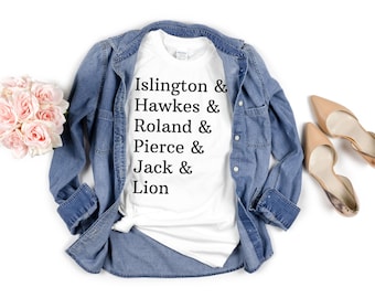 Può includere: Maglietta bianca con la scritta "Islington & Hawkes & Roland & Pierce & Jack & Lion" stampata su di essa. La maglietta è appoggiata su una superficie bianca con una camicia di jeans e un paio di scarpe beige.