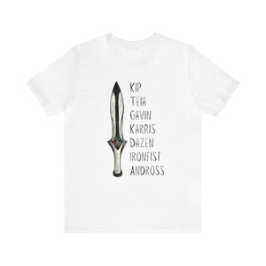 May include: White t-shirt featuring a stylized dagger design with a colorful gem-like detail. The names "Kip, Teia, Gavin, Karris, Dazen, Ironfist, Andross" are printed vertically next to the dagger. The shirt is made of a soft, lightweight fabric.