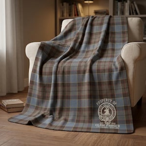 May include: A soft throw blanket featuring a blue, brown, and grey plaid pattern. The lower right corner displays a circular crest with a stag head, the text "PROPERTY OF CLAN FRASER", "JE SUIS PREST", "EST 1743", and "LALLYBROCH - SCOTLAND" printed in white.