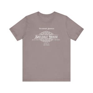 K&ouml;nnte beinhalten: Ein heidefarbenes T-Shirt mit einem wei&szlig;en Grafikdesign, auf dem "Veterinary Services Skeldale House" mit einem dekorativen Schn&ouml;rkel steht. Der Text unter der Grafik lautet "Antiquated Tradition - James Harrison - Downworthy 1921 - Braith, Yorkshire."
