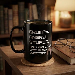 Può includere: Una tazza in ceramica nera con testo bianco che recita: GRUMPY. ANGRY. STUPID. HOW LONG SINCE LAST SLEEP QUESTION? La tazza ha un manico curvo e contiene un liquido scuro con vapore visibile che sale dall'apertura superiore del contenitore.