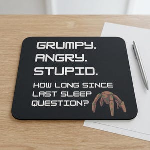 May include: A black rectangular mouse pad featuring white text that reads: GRUMPY. ANGRY. STUPID. HOW LONG SINCE LAST SLEEP QUESTION? A small, brown, four-legged robotic creature illustration is positioned in the bottom right corner of the pad.