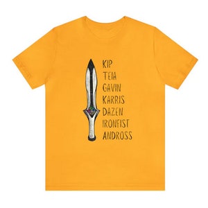 May include: Mustard yellow t-shirt featuring a stylized dagger illustration with the names Kip, Teia, Gavin, Karris, Dazen, Ironfist, and Andross printed vertically. The dagger has a black blade and a silver handle with colorful accents.