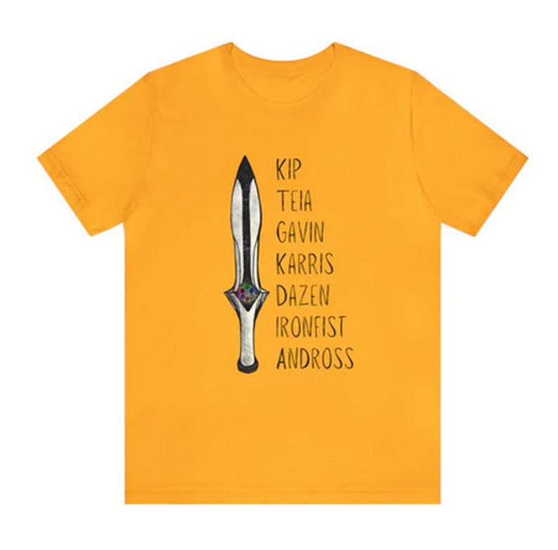 May include: Mustard yellow t-shirt featuring a stylized dagger illustration with the names Kip, Teia, Gavin, Karris, Dazen, Ironfist, and Andross printed vertically. The dagger has a black blade and a silver handle with colorful accents.