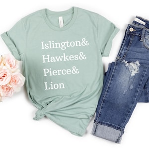 May include: A sage green short-sleeve t-shirt with white text reading Islington& Hawkes& Pierce& Lion, paired with dark blue distressed denim jeans and a cluster of light pink artificial roses.