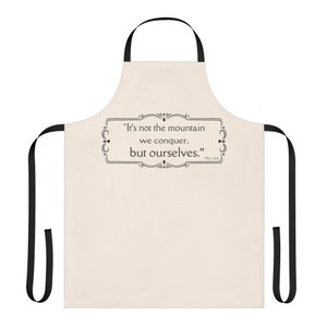 May include: A beige kitchen apron with black straps. The apron has a quote printed on it: "It's not the mountain we conquer, but ourselves." -  Ed  Hale