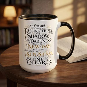 May include: A white ceramic mug with a black interior and handle, featuring black and gold decorative typography that reads: In the end, it's only a passing thing, this shadow. Even darkness must pass. A new day will come. And when the sun shines, it'll shine out the clearer.