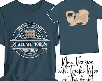 Könnte beinhalten: Ein dunkelblaues T-Shirt mit einem weißen Kreisdesign mit dem Text "Farnon & Herriot Skeldale House Darrowby 2297". Die Rückseite des Shirts zeigt eine Cartoon-Illustration eines hellbraunen Hundes mit schwarzen Augen und einer schwarzen Nase.