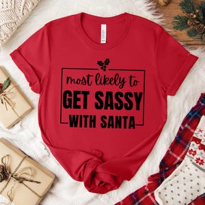 Può includere: Una maglietta rossa con la scritta nera "most likely to GET SASSY WITH SANTA". Il design include un rametto di agrifoglio. La maglietta ha un girocollo e maniche arrotolate. Abbigliamento a tema natalizio.