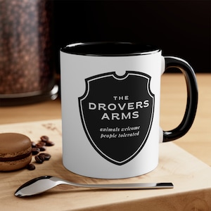 May include: White ceramic mug with a black rim and handle. The mug features a black and white shield with the text "The Drovers Arms" and the text "animals welcome people tolerated".
