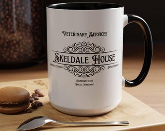 May include: A white ceramic mug with a black interior and handle. The front features black text reading Veterinary Services, Skeldale House, Siegfried Farnon, James Herriot, Darrowby 2297, and Dales, Yorkshire, surrounded by decorative scrollwork.