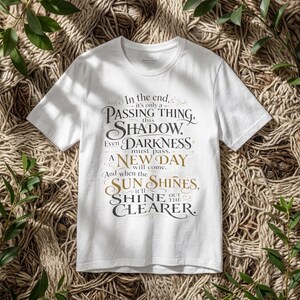 Op de afbeelding: Een wit T-shirt met korte mouwen en zwarte en gouden decoratieve typografie met de tekst: In the end, it's only a PASSING THING, this SHADOW. Even DARKNESS must pass. A NEW DAY will come. And when the SUN SHINES, it'll SHINE out the CLEARER.