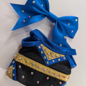 K&ouml;nnte beinhalten: Eine blaue Cheerleader-Schleife und ein passendes schwarz-goldenes Uniformteil. Die Schleife besteht aus einem gl&auml;nzenden blauen Stoff und ist mit schillernden Strasssteinen verziert. Das Uniformteil ist schwarz mit goldenen Akzenten und Strasssteinen.