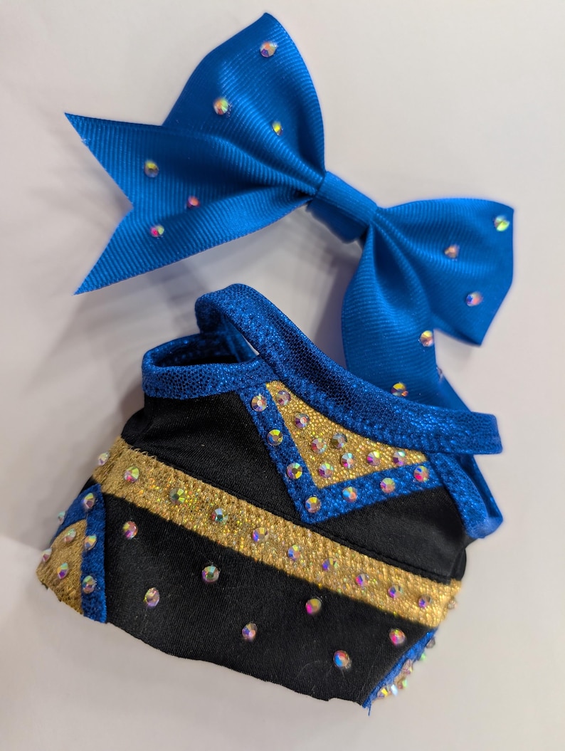K&ouml;nnte beinhalten: Eine blaue Cheerleader-Schleife und ein passendes schwarz-goldenes Uniformteil. Die Schleife besteht aus einem gl&auml;nzenden blauen Stoff und ist mit schillernden Strasssteinen verziert. Das Uniformteil ist schwarz mit goldenen Akzenten und Strasssteinen.
