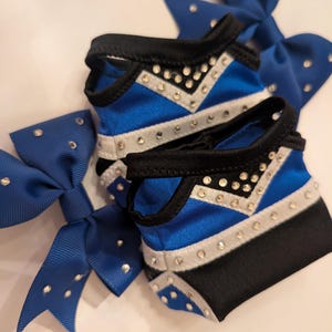 K&ouml;nnte beinhalten: Nahaufnahme von Cheerleading-Zubeh&ouml;r. Zwei blau-schwarze Cheerleading-Schleifen mit silbernen Strasssteinen liegen neben zwei passenden Armb&auml;ndern. Die Armb&auml;nder haben ein blau-silbernes Chevron-Design mit Strasssteinen. Die Schleifen sind aus gl&auml;nzendem blauem Band gefertigt.