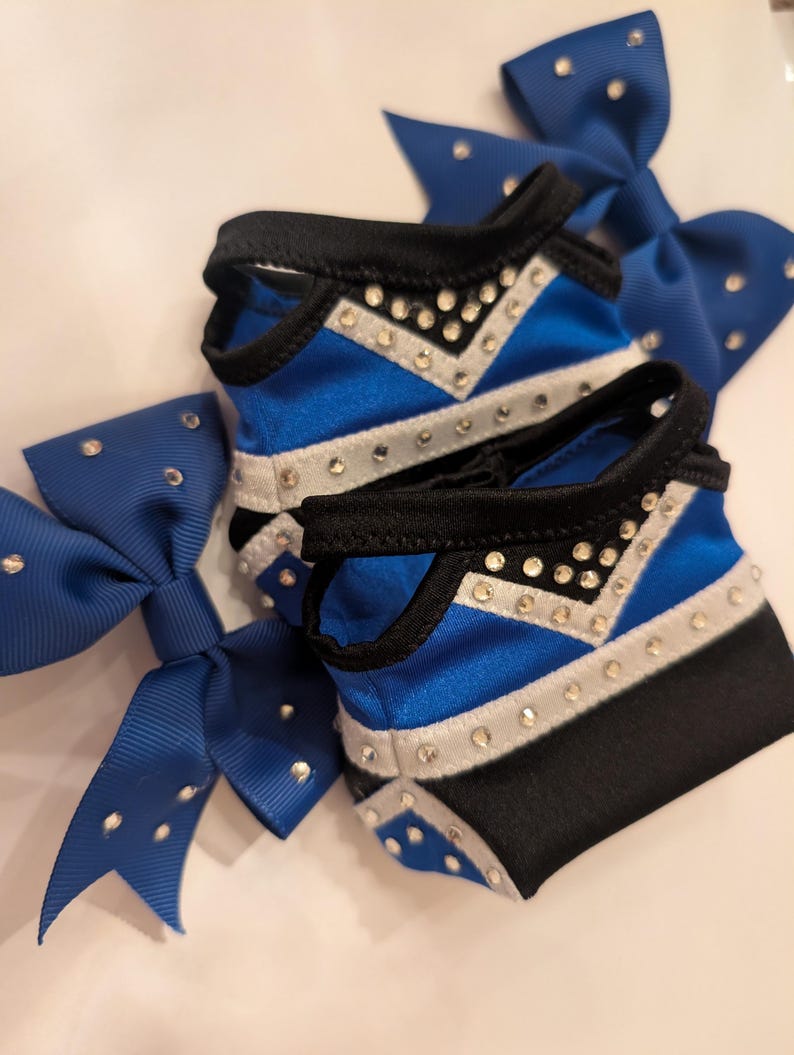 K&ouml;nnte beinhalten: Nahaufnahme von Cheerleading-Zubeh&ouml;r. Zwei blau-schwarze Cheerleading-Schleifen mit silbernen Strasssteinen liegen neben zwei passenden Armb&auml;ndern. Die Armb&auml;nder haben ein blau-silbernes Chevron-Design mit Strasssteinen. Die Schleifen sind aus gl&auml;nzendem blauem Band gefertigt.