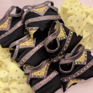 K&ouml;nnte beinhalten: Eine Sammlung schwarzer und gelber Cheerleading-Accessoires. Die Accessoires haben silberne Verzierungen und sind mit Strasssteinen verziert. Mehrere gelbe Schleifen mit Strasssteinen sind ebenfalls sichtbar.