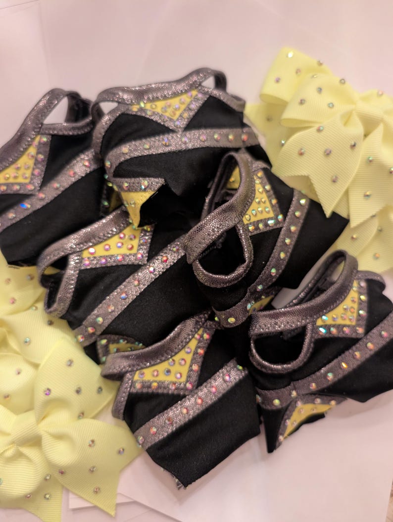 K&ouml;nnte beinhalten: Eine Sammlung schwarzer und gelber Cheerleading-Accessoires. Die Accessoires haben silberne Verzierungen und sind mit Strasssteinen verziert. Mehrere gelbe Schleifen mit Strasssteinen sind ebenfalls sichtbar.