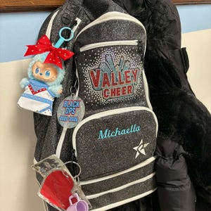 K&ouml;nnte beinhalten: Ein schwarzer Glitzer-Rucksack mit wei&szlig;em Besatz und dem Text "VALLEY CHEER" in roten und blauen Strasssteinen. Der Name "Michaella" ist t&uuml;rkis gestickt. Eine blaue Puppe mit roter Schleife und Schl&uuml;sselanh&auml;nger h&auml;ngen am Rucksack.