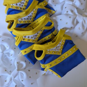 K&ouml;nnte beinhalten: Nahaufnahme von Cheerleading-Zubeh&ouml;r. Mehrere blau-gelbe Cheerleading-Oberteile mit Strassverzierungen sind aufeinander gestapelt. Wei&szlig;e Schleifen mit Strasssteinen sind ebenfalls sichtbar.