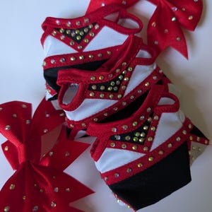 K&ouml;nnte beinhalten: Mehrere Cheerleader-Schleifen und passende Uniformteile. Die Schleifen sind rot mit verstreuten Strasssteinen. Die Uniformteile haben wei&szlig;e, rote und schwarze Akzente, ebenfalls mit Strasssteinen verziert. Die Artikel sind auf einer wei&szlig;en Oberfl&auml;che angeordnet.