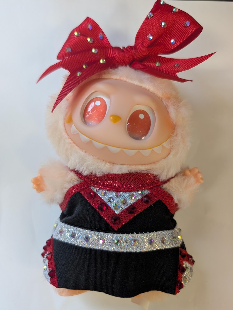 K&ouml;nnte beinhalten: Eine skurrile Puppe mit pfirsichfarbenem Gesicht, gro&szlig;en roten Augen und einem grinsenden Mund. Sie tr&auml;gt ein schwarz-rotes Kleid, das mit Strasssteinen und einer gro&szlig;en roten Schleife mit glitzernden Akzenten verziert ist. Die Puppe hat eine flauschige, hellfarbige Kapuze.