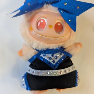 K&ouml;nnte beinhalten: Eine Pl&uuml;schpuppe mit hellpfirsichfarbenem Gesicht, roten Augen und einem grinsenden Mund. Sie tr&auml;gt ein schwarz-blaues Outfit mit Strassakzenten und eine gro&szlig;e blaue Schleife mit Strasssteinen. Die Puppe hat einen flauschigen, hellpfirsichfarbenen Fellbesatz um ihr Gesicht.
