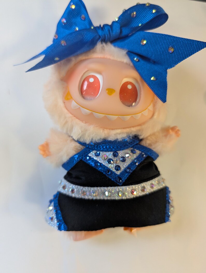 K&ouml;nnte beinhalten: Eine Pl&uuml;schpuppe mit hellpfirsichfarbenem Gesicht, roten Augen und einem grinsenden Mund. Sie tr&auml;gt ein schwarz-blaues Outfit mit Strassakzenten und eine gro&szlig;e blaue Schleife mit Strasssteinen. Die Puppe hat einen flauschigen, hellpfirsichfarbenen Fellbesatz um ihr Gesicht.
