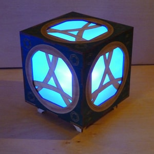 May include: A blue and green wooden cube with a glowing blue light inside. The cube has a geometric design on the outside and is lit from within.