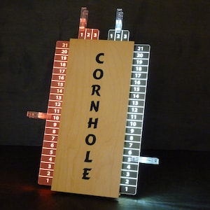 May include: A wooden cornhole game set with two scorekeeping boards. Each board has a white or red plastic clip that slides up and down to keep score. The boards are numbered 1 to 21. The center of the boards has the word "CORNHOLE" printed in black.