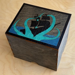 May include: A wooden box with a nautical design. The top features a black silhouette of a ship, teal tentacles, and blue waves. The box is square-shaped with a dark gray finish.