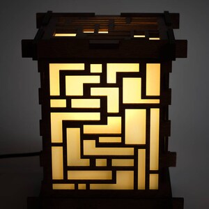 May include: A wooden lamp with a geometric design. The lamp is lit and the light shines through the cutouts in the wood. The lamp is on a dark background.