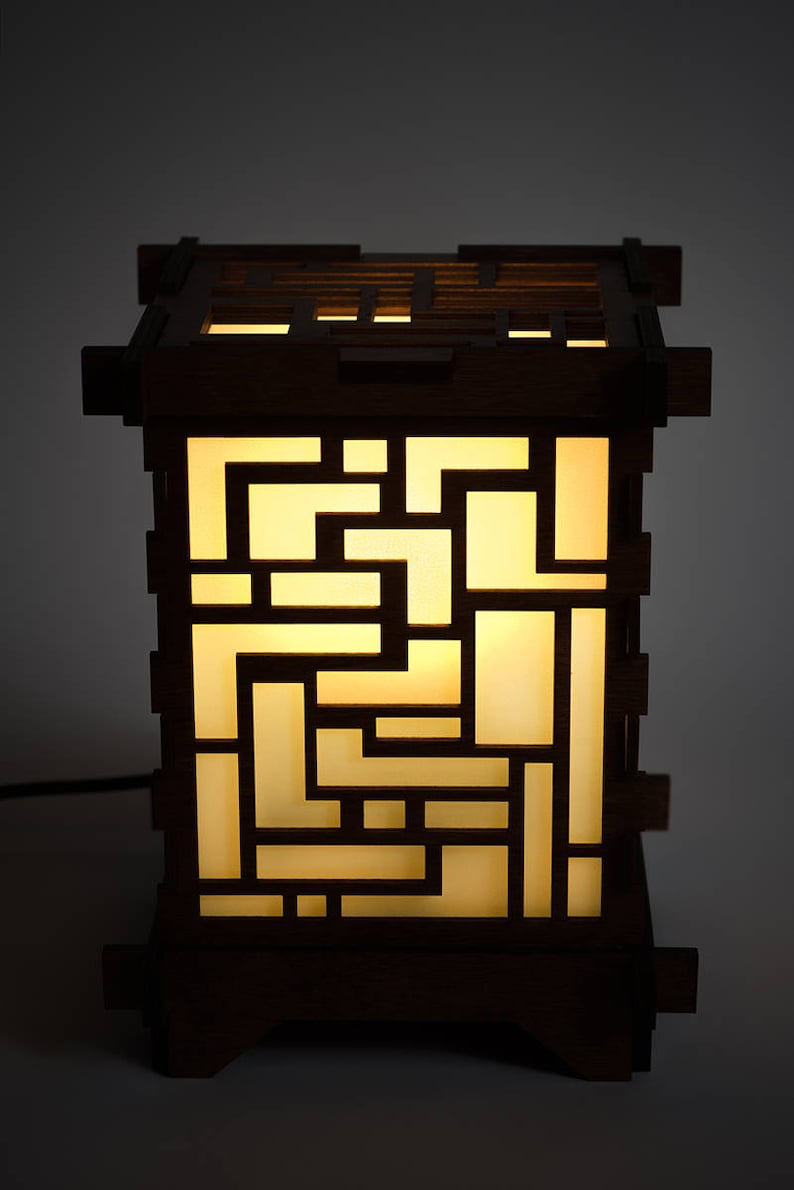 May include: A wooden lamp with a geometric design. The lamp is lit and the light shines through the cutouts in the wood. The lamp is on a dark background.