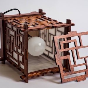 May include: A wooden lamp with a geometric design. The lamp has a light bulb inside and a removable panel. The lamp is made of dark wood and has a white interior.