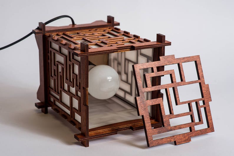 May include: A wooden lamp with a geometric design. The lamp has a light bulb inside and a removable panel. The lamp is made of dark wood and has a white interior.