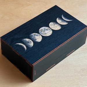 Puede incluir: Caja rectangular de madera negra con tapa azul oscuro decorada con fases lunares blancas y beige. La caja está sobre una superficie de madera marrón claro.