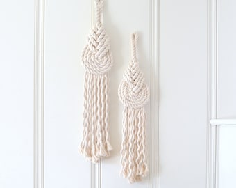 May include: Two macrame wall hangings in a cream color, with a knotted design. The hangings are displayed on a white wall with a wood paneling effect. The hangings are different sizes, with the larger one on the left and the smaller one on the right.