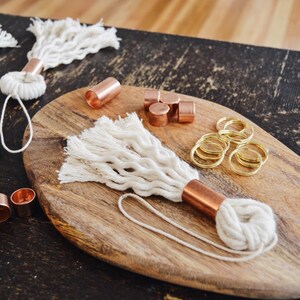 May include: A close-up of a wooden cutting board with a white rope tassel keychain, copper end caps, and gold jump rings. There are also several copper end caps and gold jump rings scattered around the board.