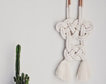 May include: A white macrame wall hanging with two large knots and two tassels. The hanging is made with natural cotton rope and has a brown accent at the top.